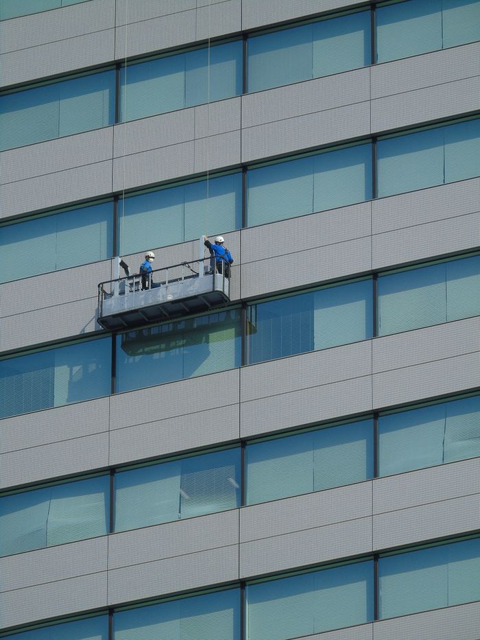 about-01 Two workers clean windows on a modern glass building facade using a suspended platform.